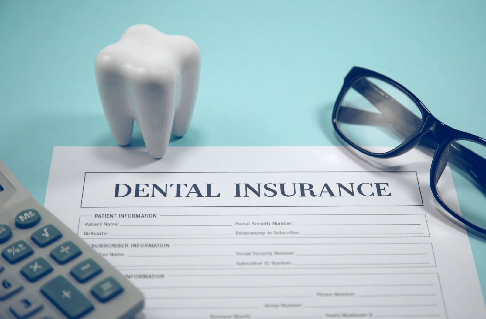 A dental insurance form sitting next to a calculator, a pair of reading glasses, and a plastic model of a tooth
