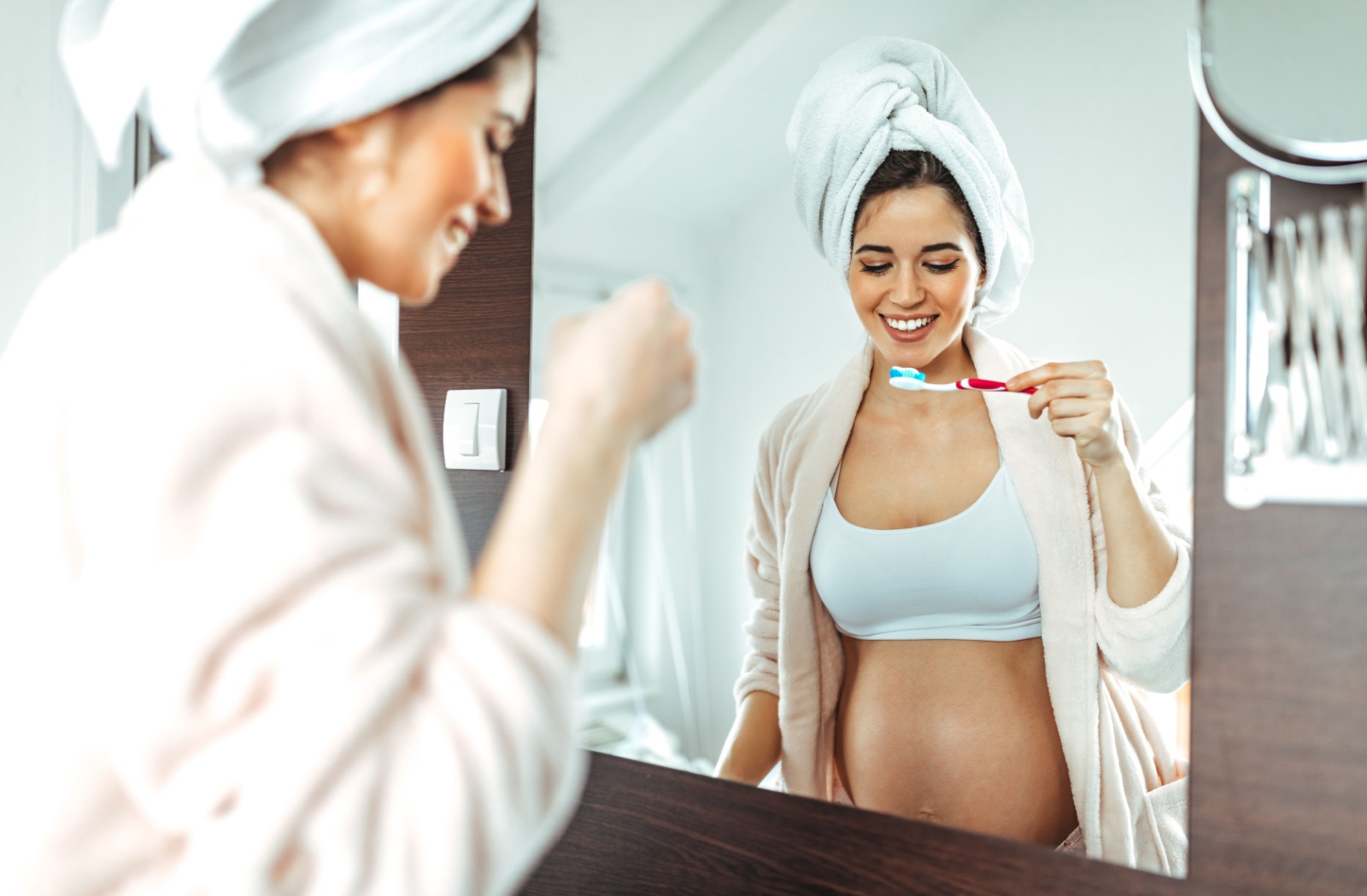 A pregnant woman smiling while brushing her teeth in front of a mirror.