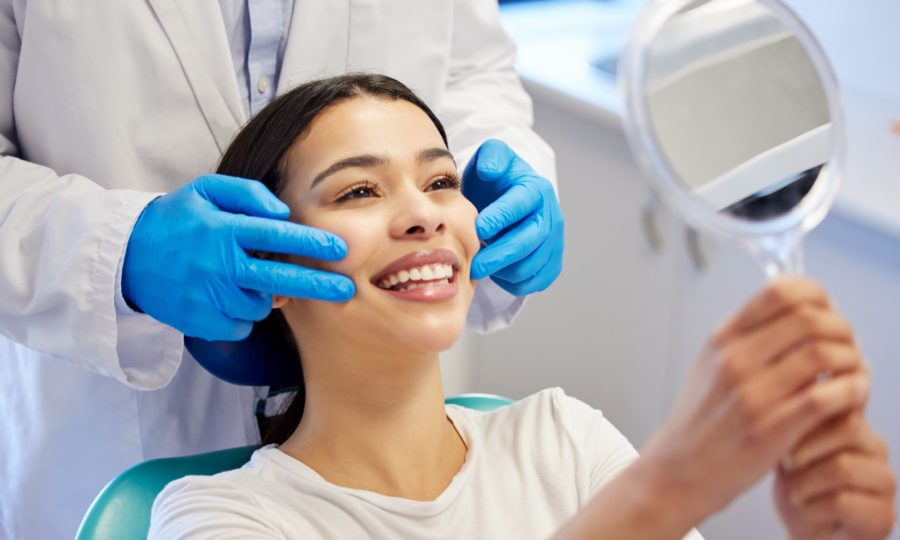 A woman smiles into a hand mirror after her teeth whitening appointment.