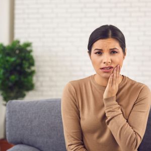 A woman sitting on the couch with her hand to one side of her jaw from tooth problems.