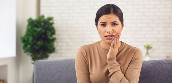 A woman sitting on the couch with her hand to one side of her jaw from tooth problems.