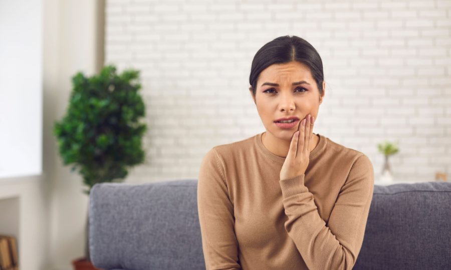 A woman sitting on the couch with her hand to one side of her jaw from tooth problems.