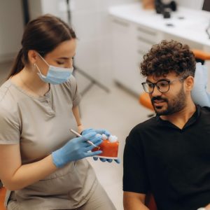 Dental professional wearing gloves and a face mask showing a model of teeth to a male patient during a consultation in a modern dental clinic.