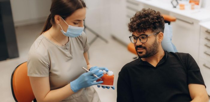 Dental professional wearing gloves and a face mask showing a model of teeth to a male patient during a consultation in a modern dental clinic.