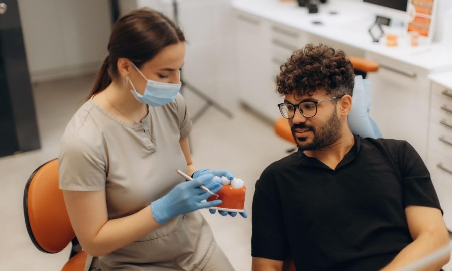 Dental professional wearing gloves and a face mask showing a model of teeth to a male patient during a consultation in a modern dental clinic.
