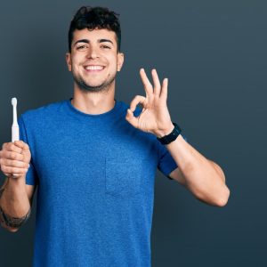 A young man holding an electric toothbrush with his right hand and doing an okay sign with his left hand