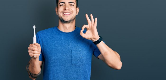 A young man holding an electric toothbrush with his right hand and doing an okay sign with his left hand