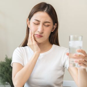 person holds a glass of ice water and rubs their jaw due to sensitive teeth