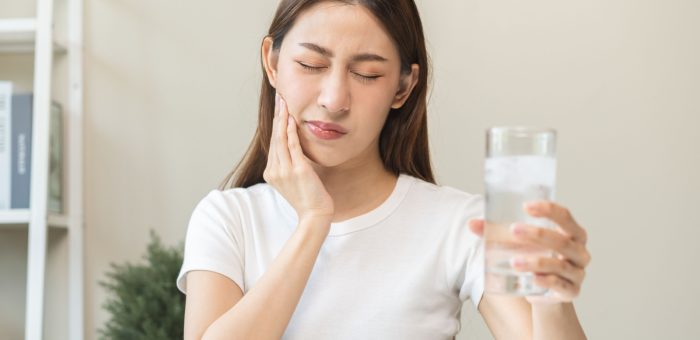 person holds a glass of ice water and rubs their jaw due to sensitive teeth