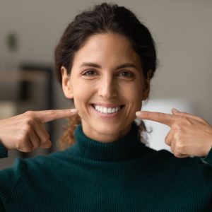 A patient smiling in their home and pointing at the results of their teeth whitening appointment.