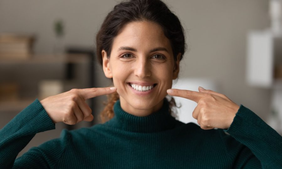 A patient smiling in their home and pointing at the results of their teeth whitening appointment.